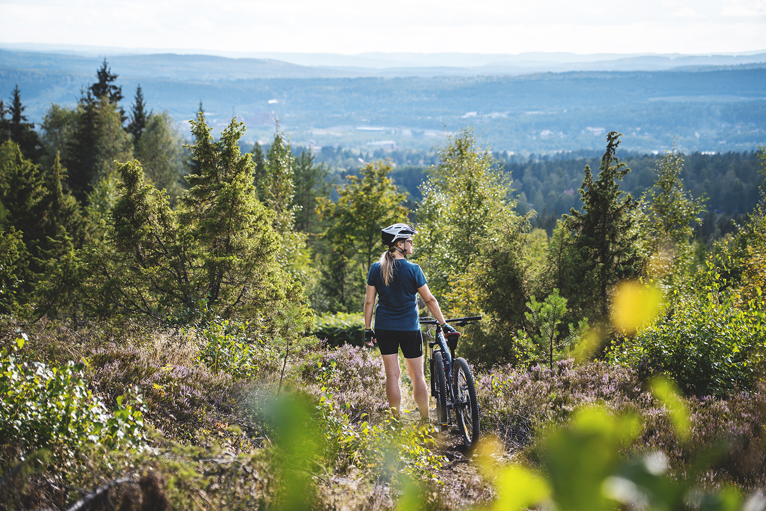 En kvinna står med en mountainbike i skogsmiljö med utsikt.