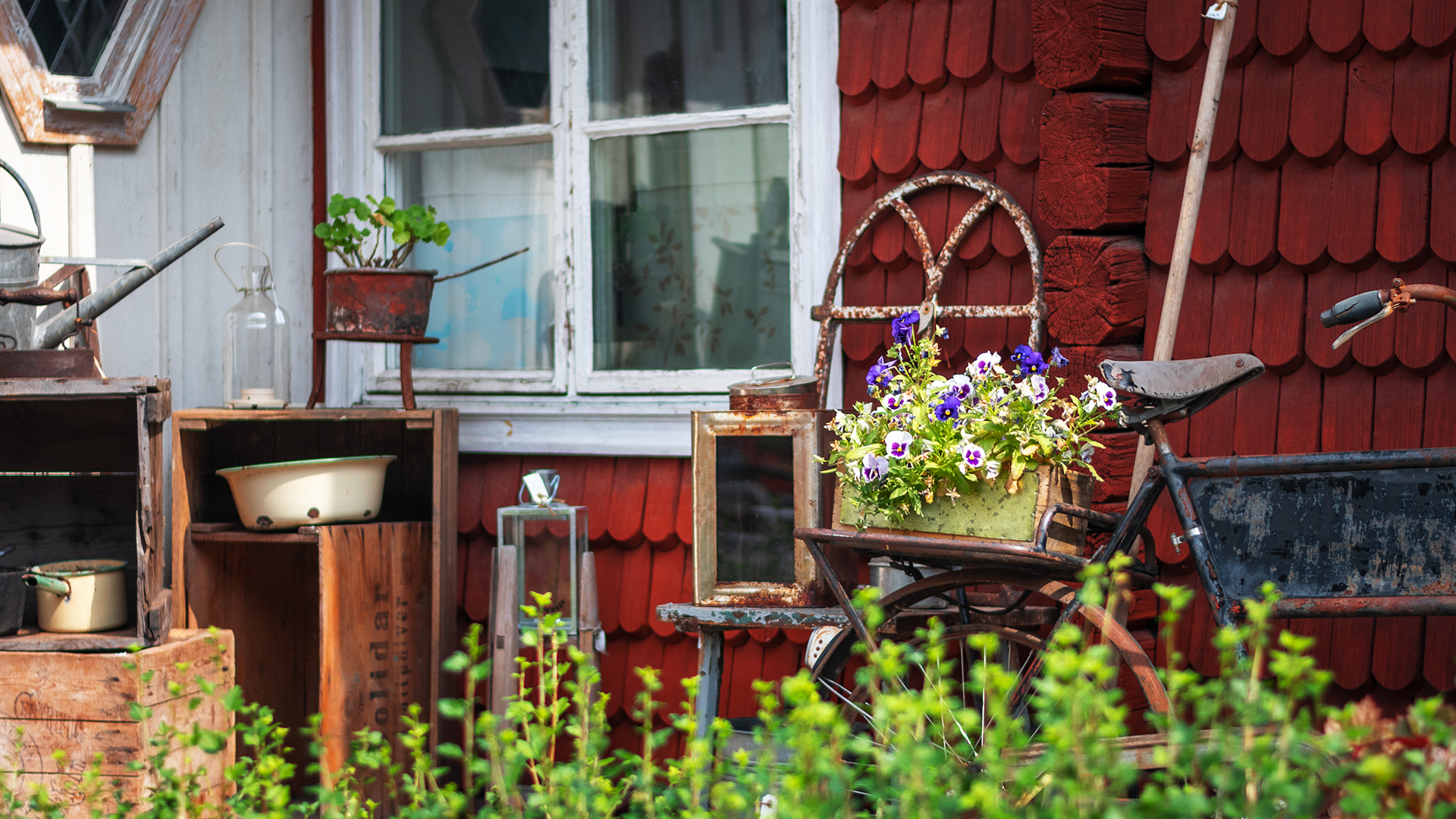 Äldre föremål och blommor framför en röd husvägg.