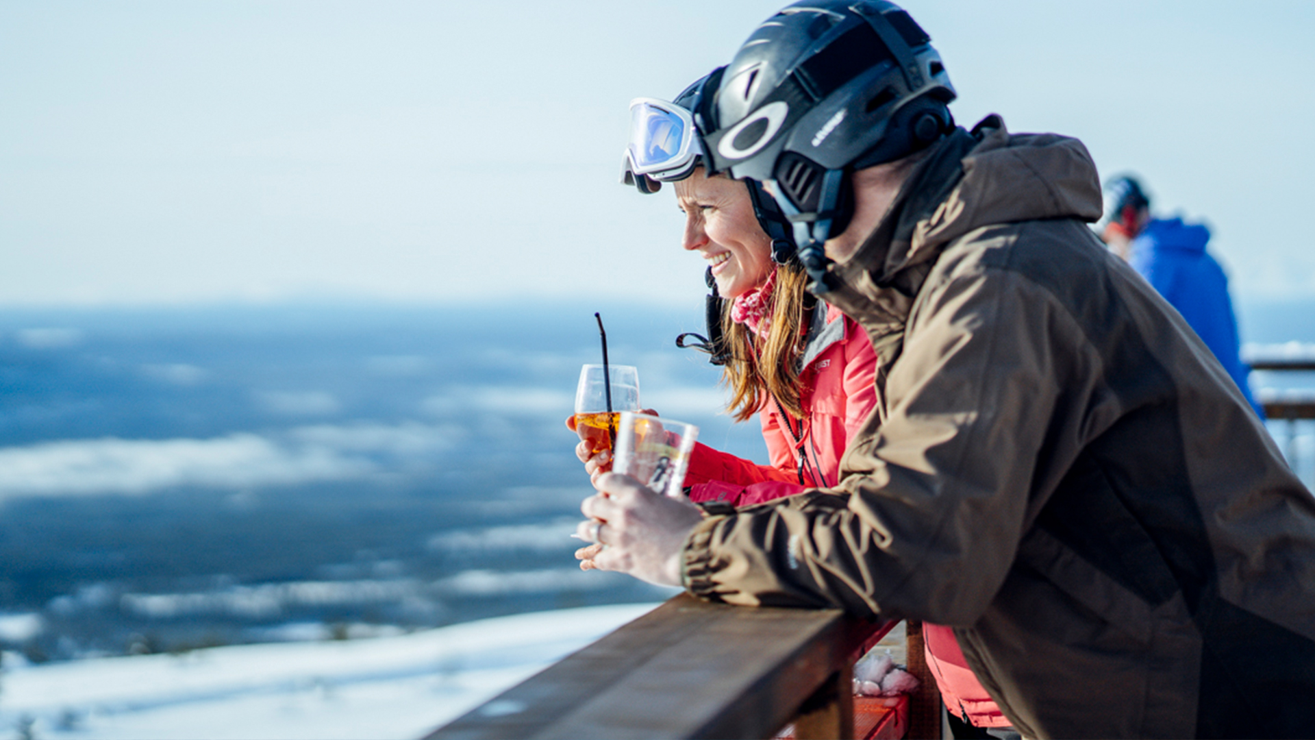 Ett par i slalomhjälmar håller i glas med dryck och lutar sig mot ett staket.