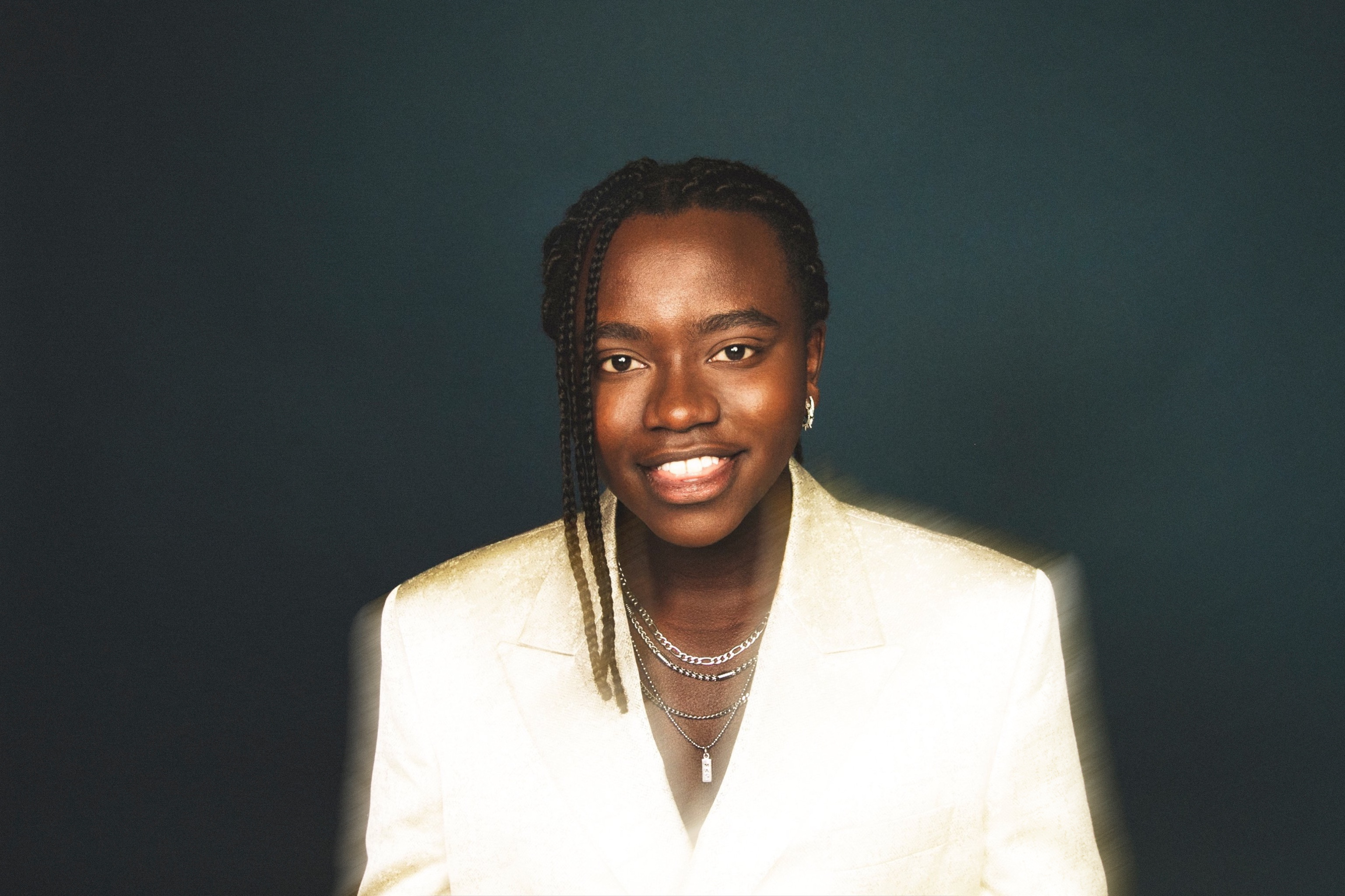 Tusse står i vit kostym mot en mörk bakgrund och ler mot kameran.