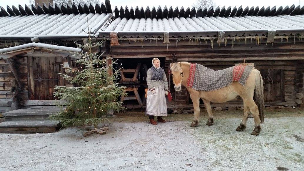 En kvinna i äldre kläder står mellan en julgran och en häst.
