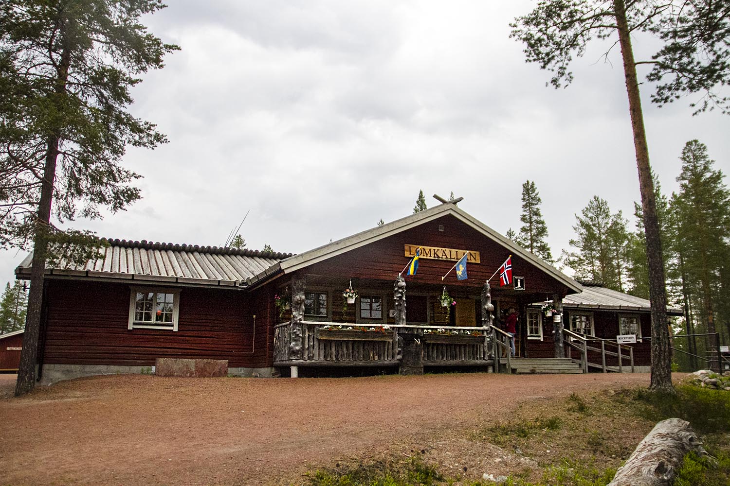 En äldre timrad byggnad med skylten Lomkällan och flaggor på fasaden.