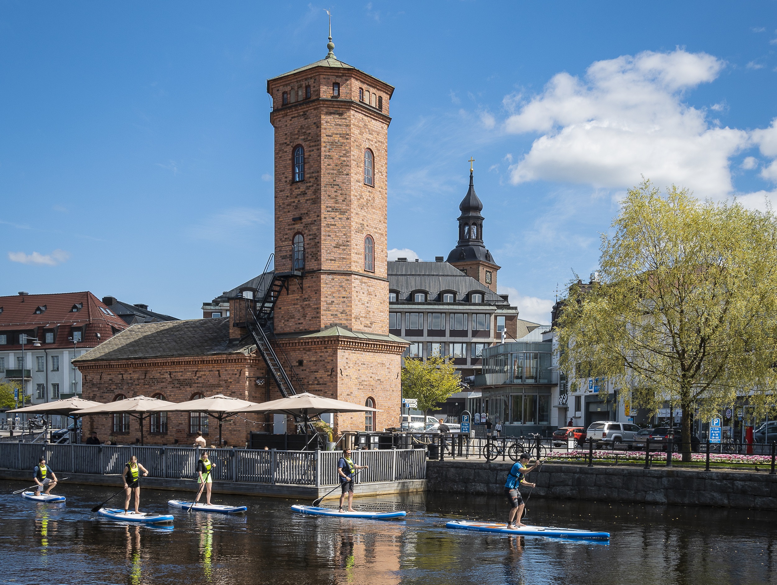 Fem personer på supbrädor som paddlar sig fram på Faluån mitt i Falu centrum förbi Spruthuset.