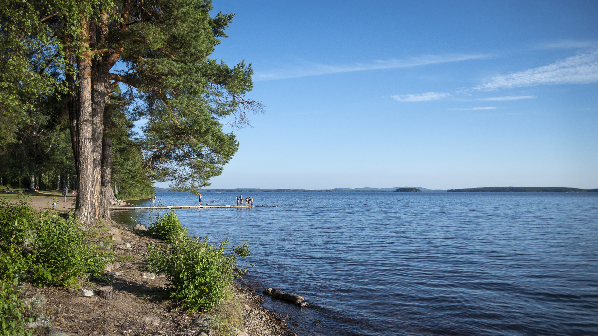 Badstrand med badande ungdomar på bryggan intill Runns härliga blåa vatten.