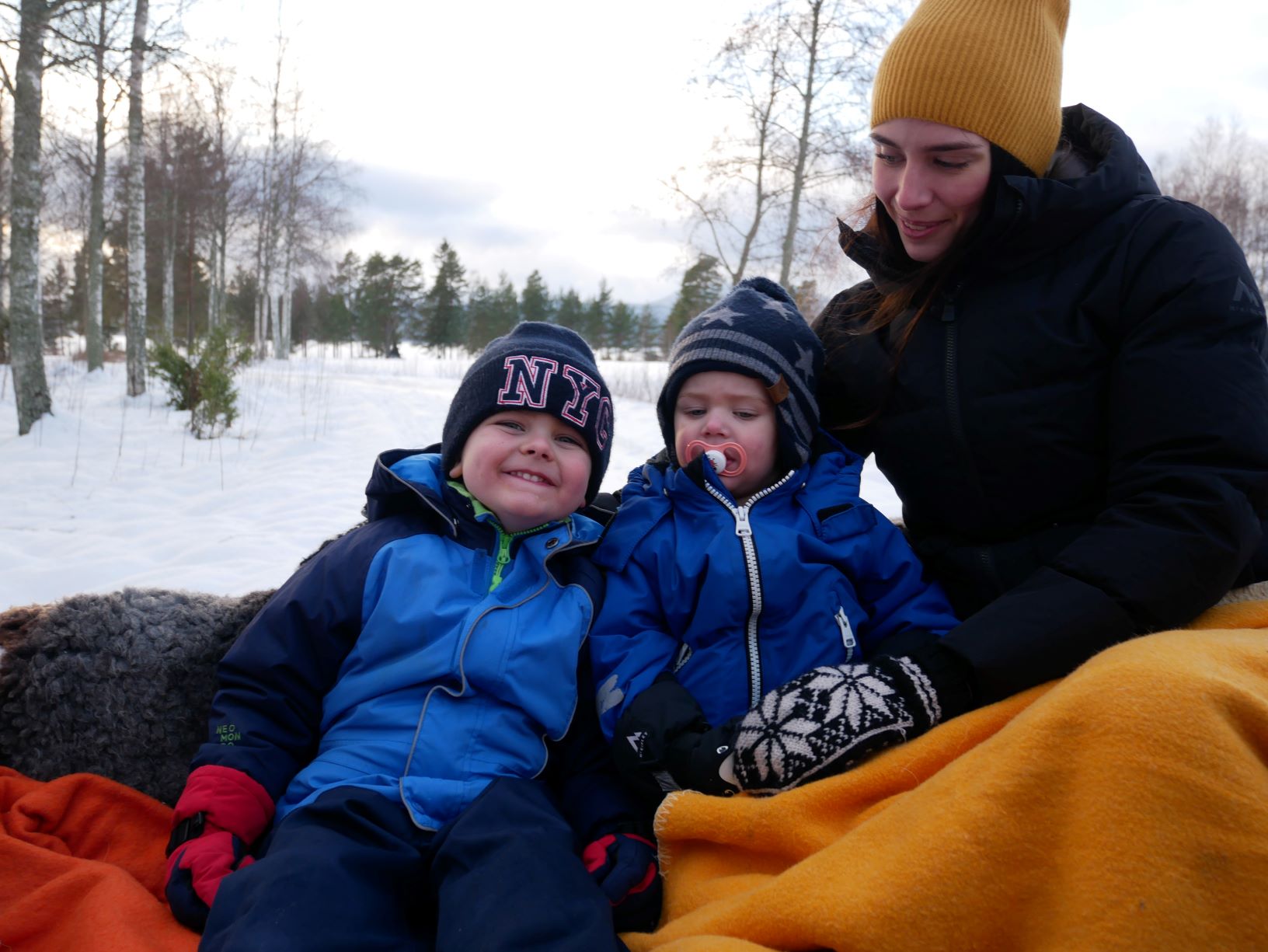 Två pojkar med mamma sitter hopkrupna i släde.