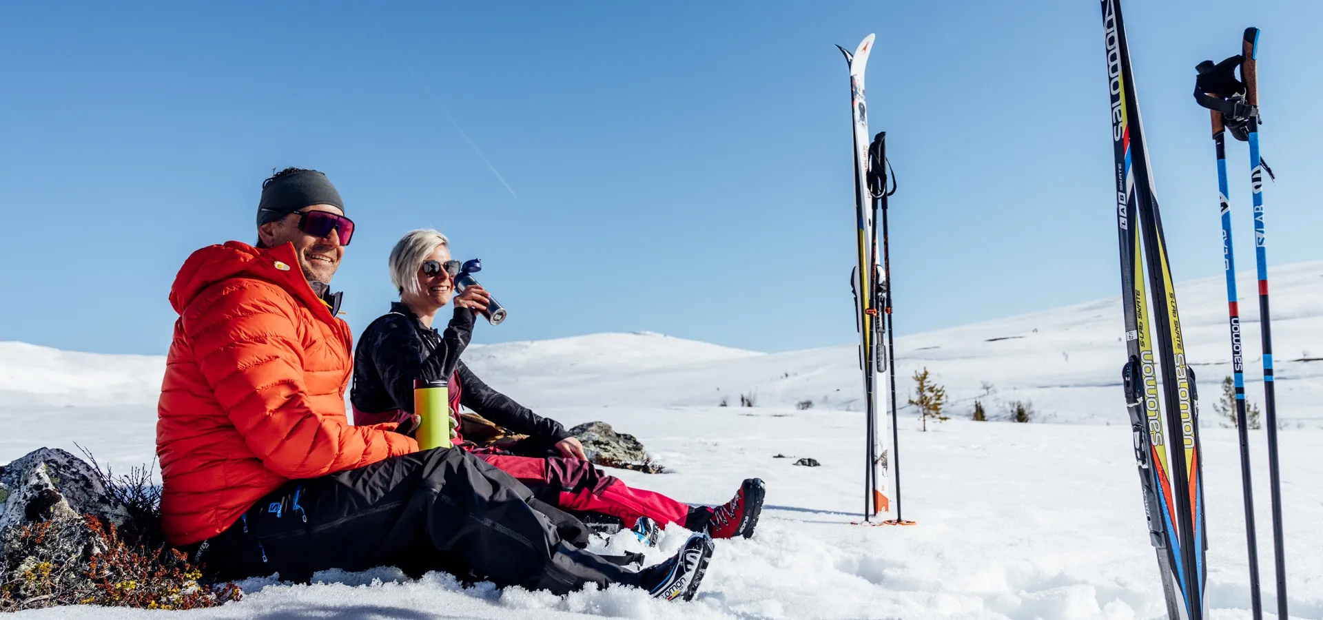 Ett par sitter och fikar i solen på fjället med skidorna nedstuckna i den djupa snön.