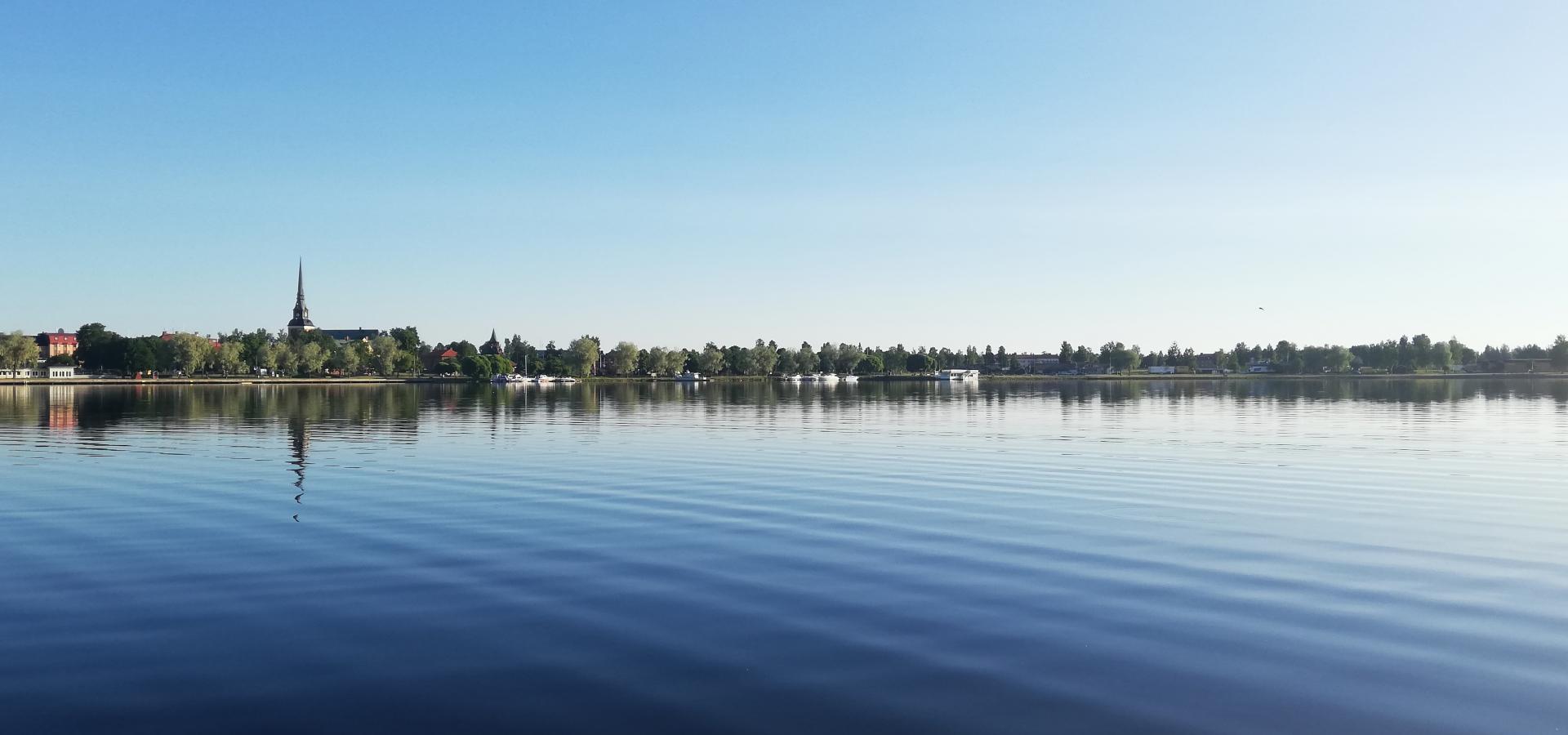 Blå himmel och blått vatten, träd och ett kyrktorn speglas i sjön.