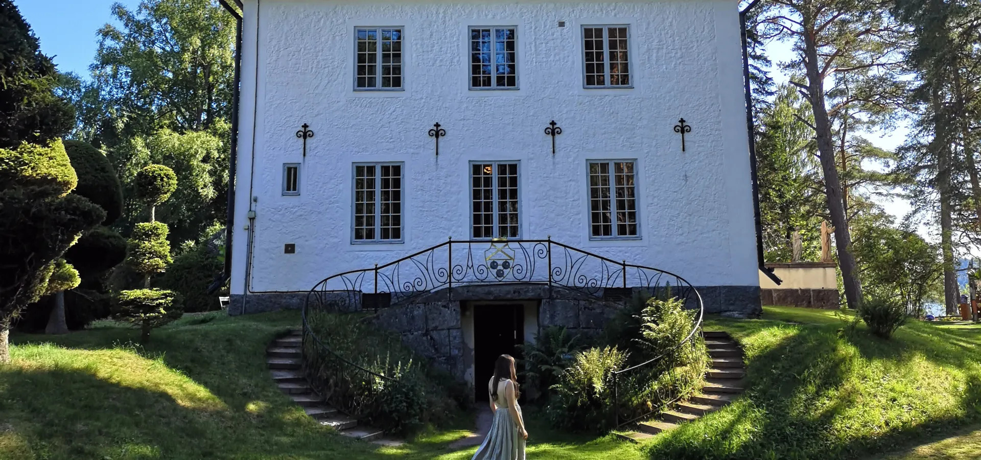En kvinna går genöm en grönskande trädgård framför ett vitt hus sommartid.