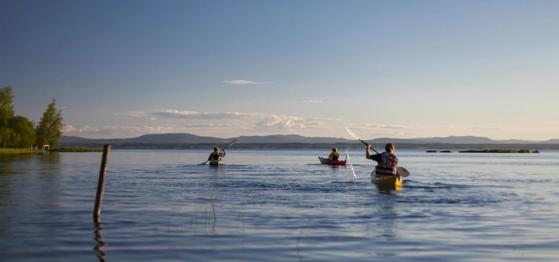 Tre personer i varsin kajak som paddlar på en sjö i skymning.