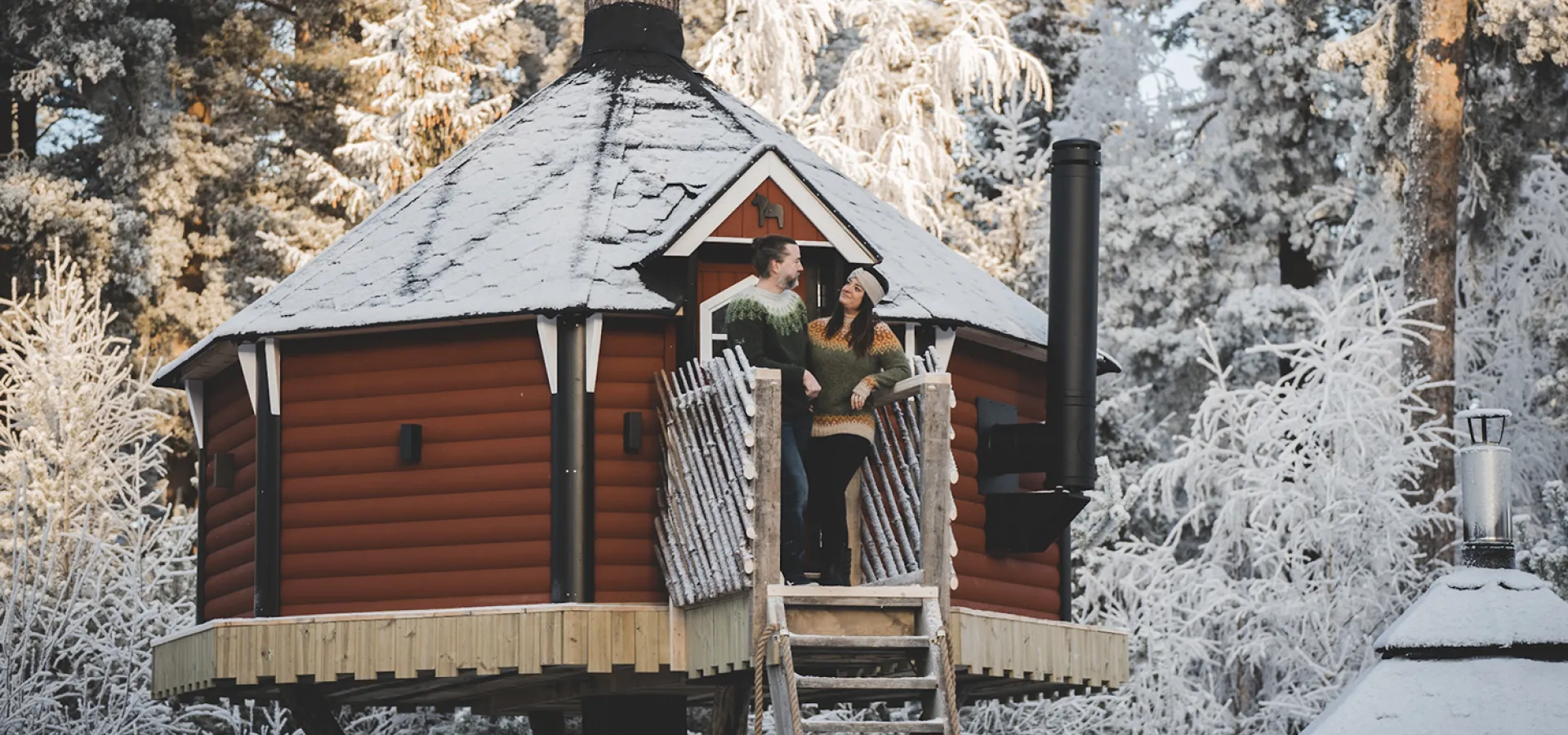 En man och en kvinna står i dörren till ett rött litet hus i ett träd.