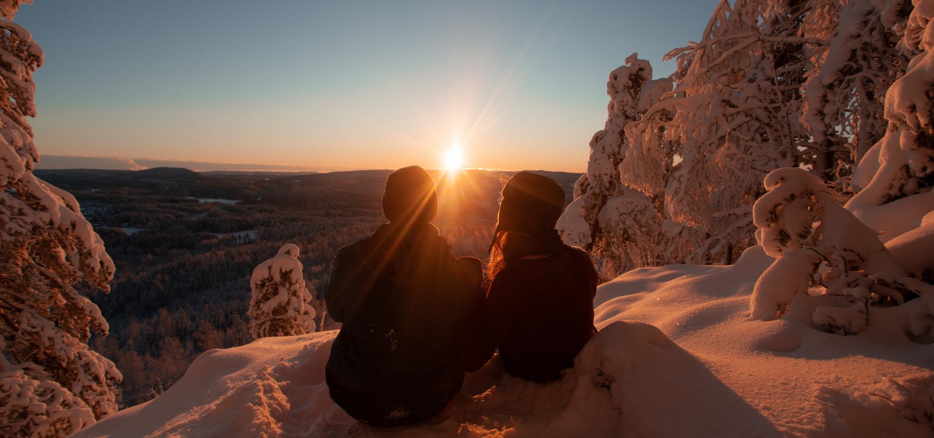 Ett par sitter i snön och ser ut över solnedgången.