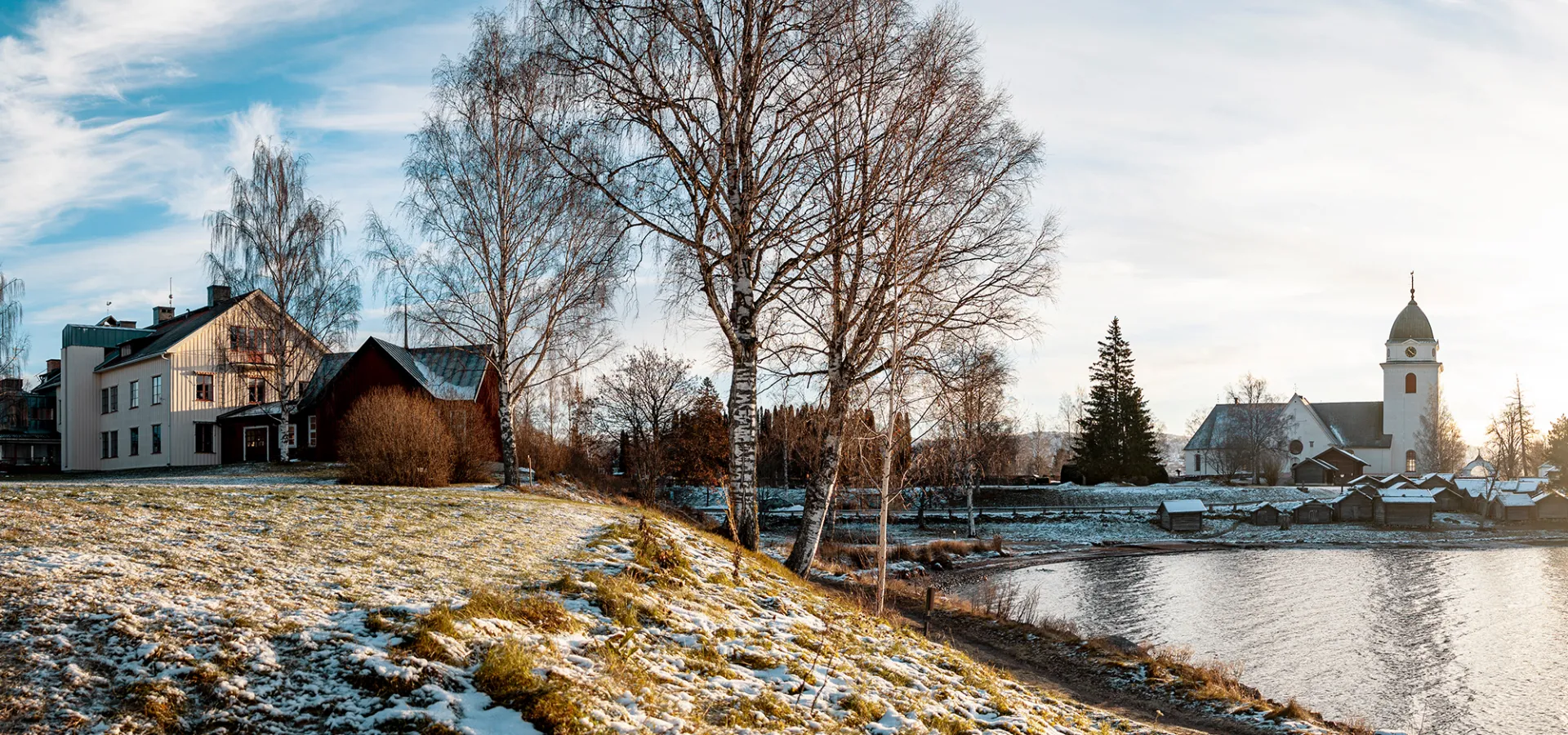 Panoramavy över ett vitt hus och en vit kyrka, gräs, träd och vatten i förgrunden.