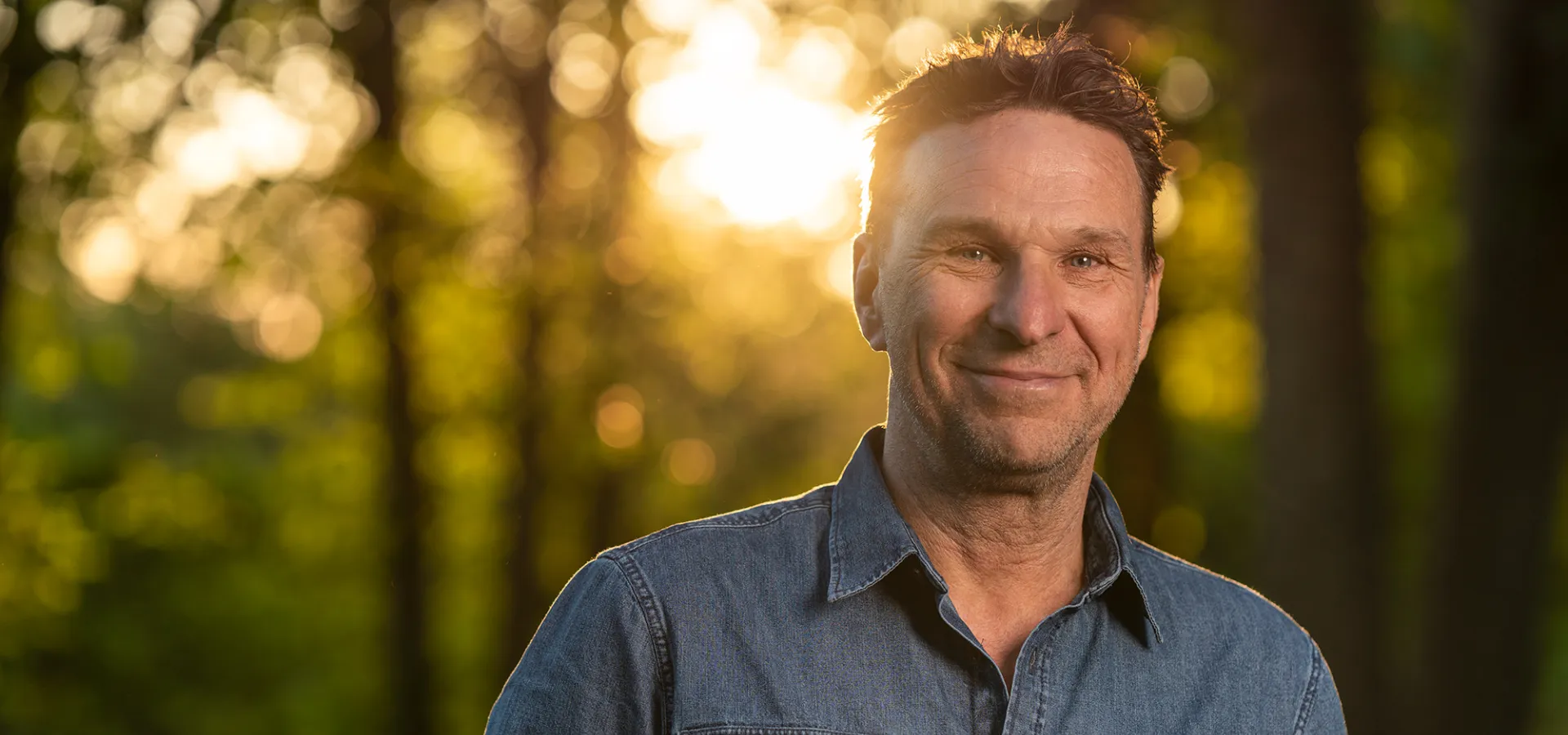 Anders Lundin ler i motljus, solen syns bakom mellan träd.