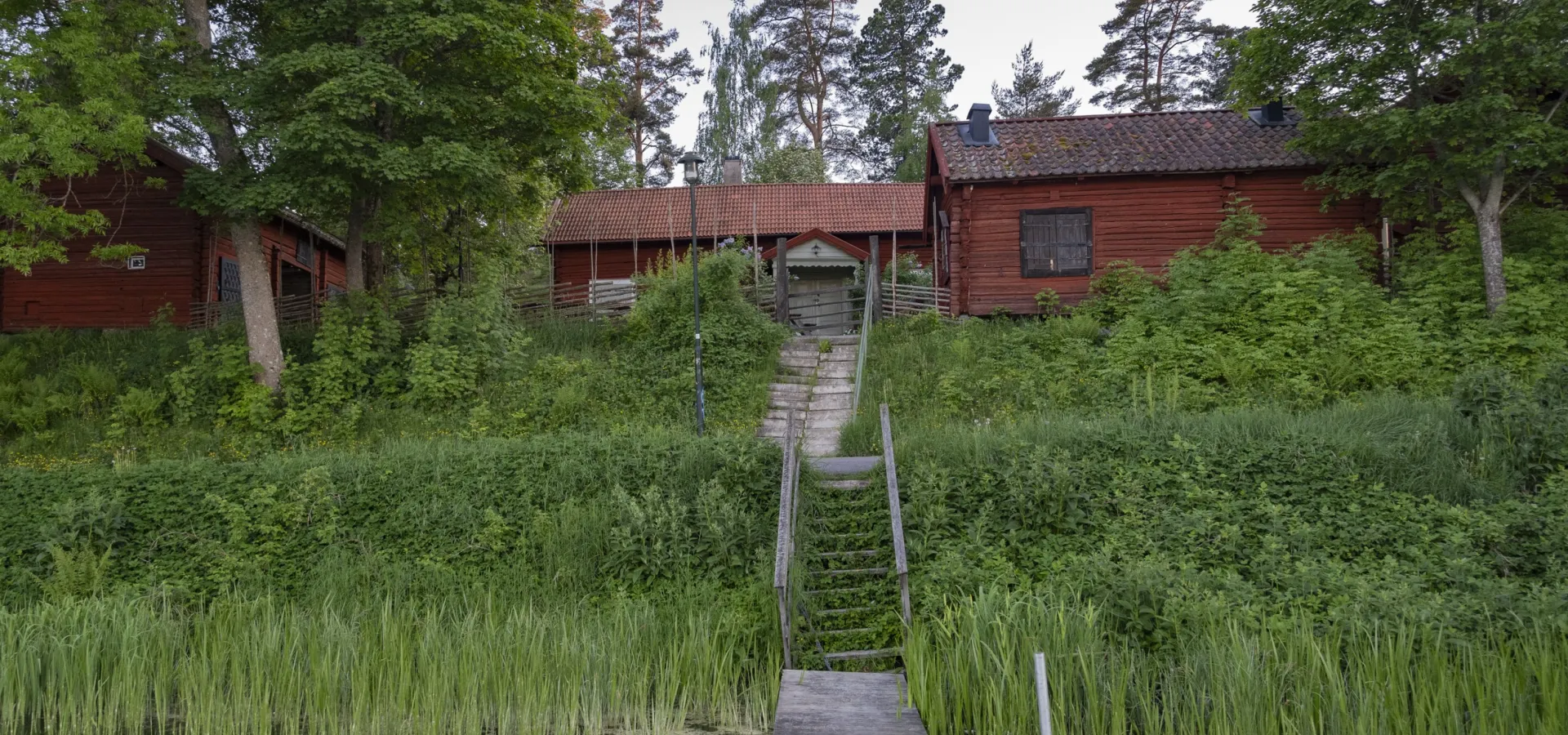Faluröda äldre hus intill en sjö omgivna av grönska.