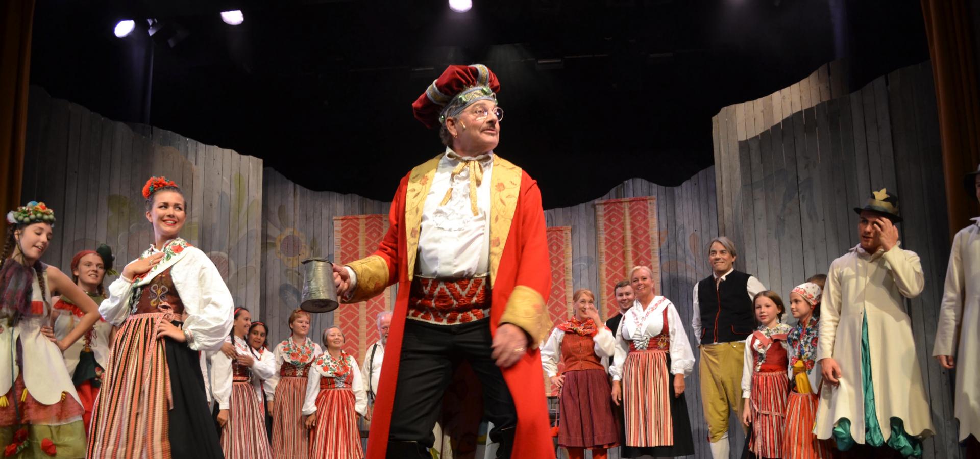 En teaterscen med människor i folkdräkt, i mitten en man i röd kappa och hatt.