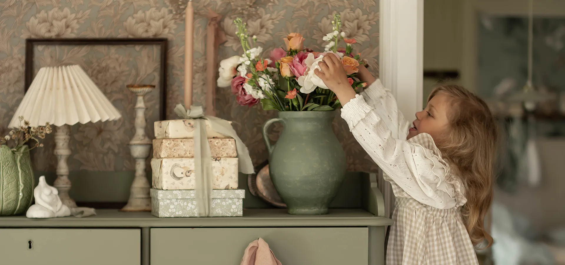 En flicka sträcker sig mot en vas med blommor som står på en grön byrå.