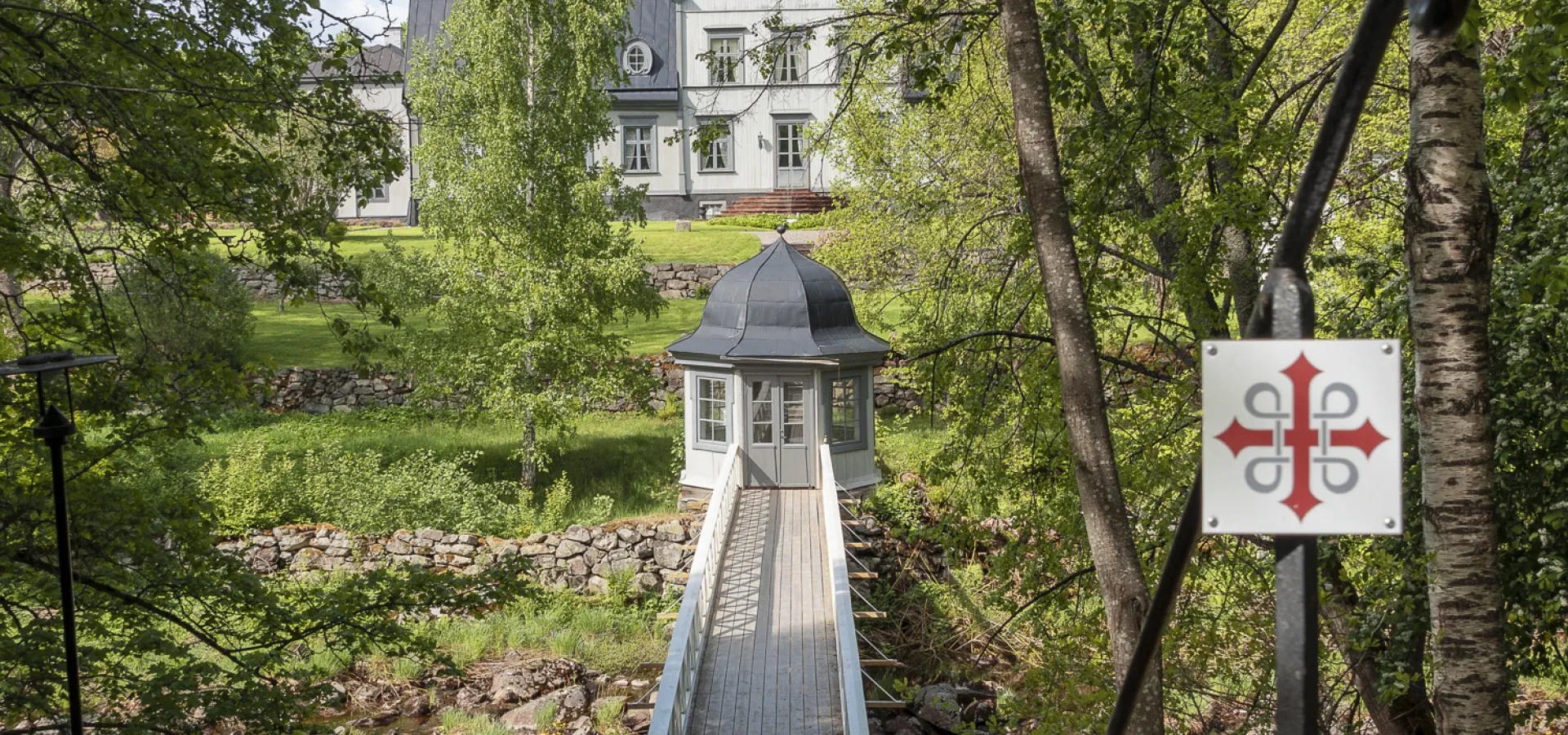 En träbro omgiven av grönska leder mot ett vitt hus.
