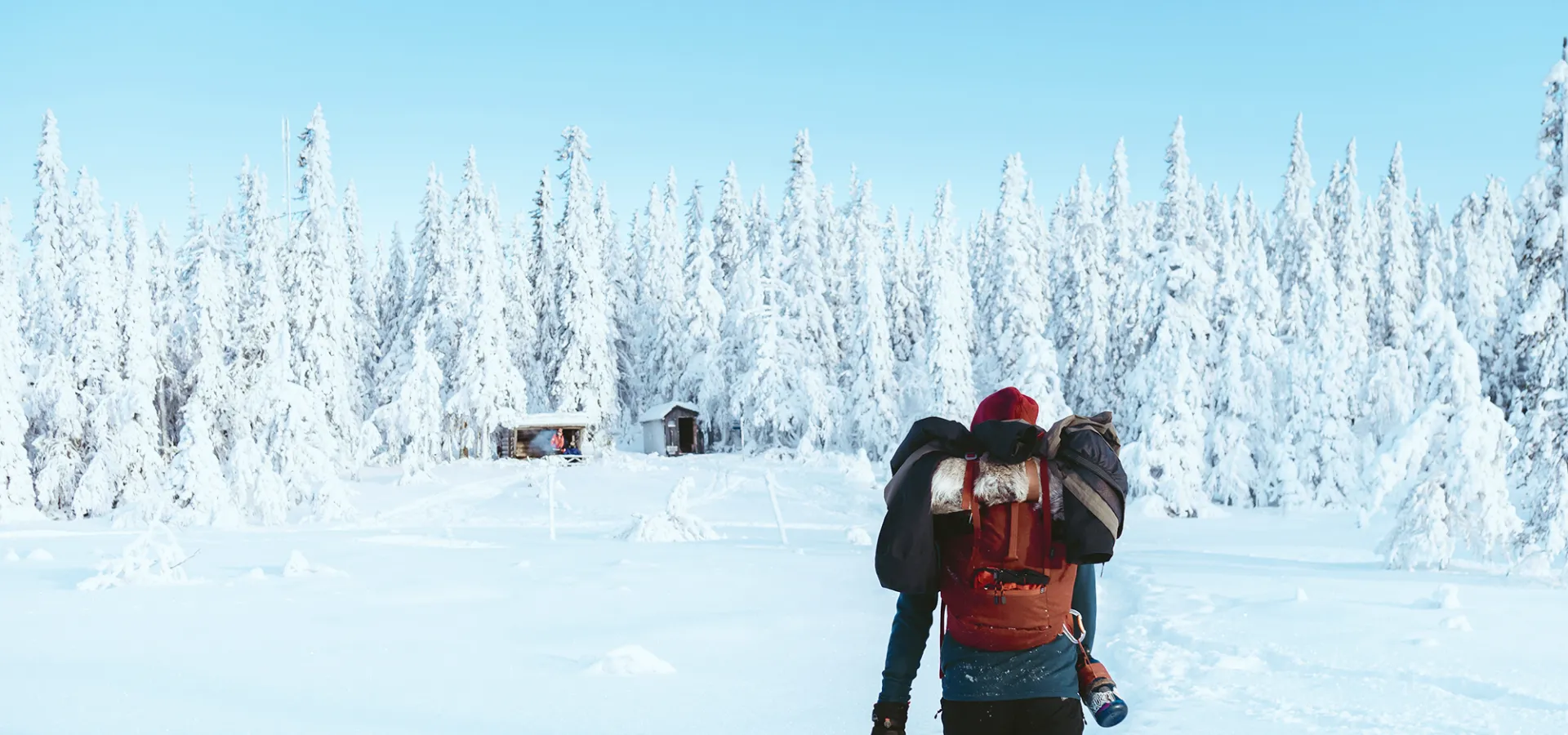 En person med ryggsäck går mot en stuga i snötäckt skog.