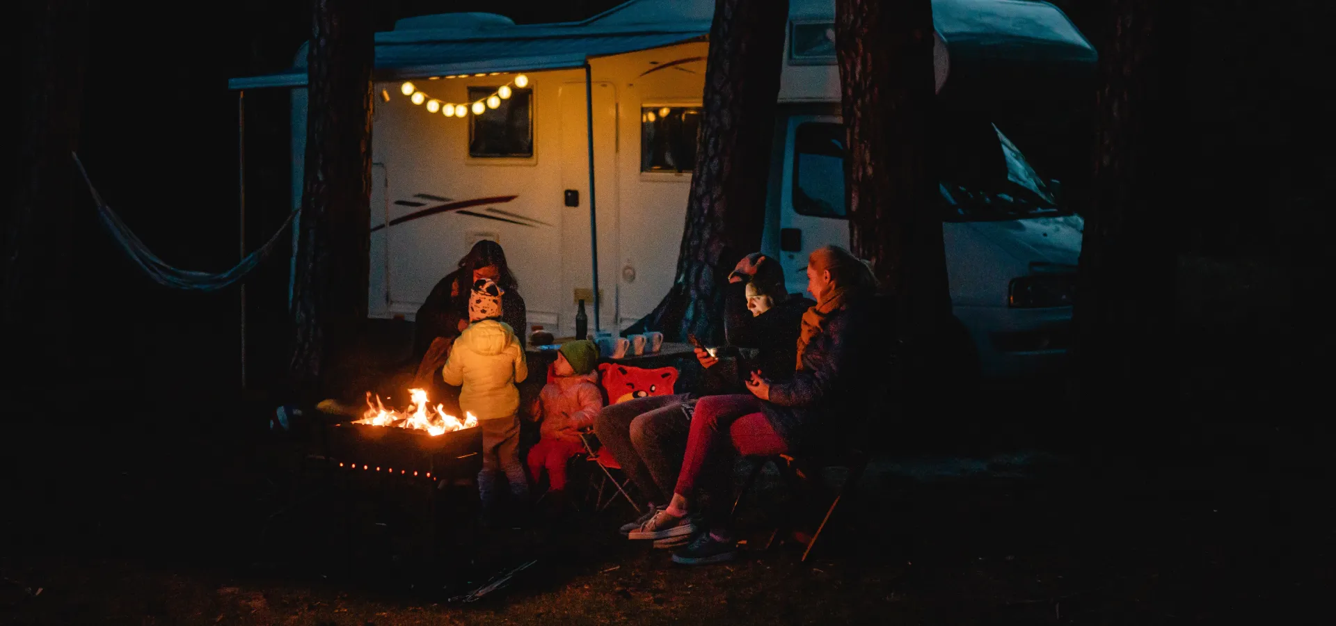 Husbil uppställd i skogen där människor sitter vid lägereld. 