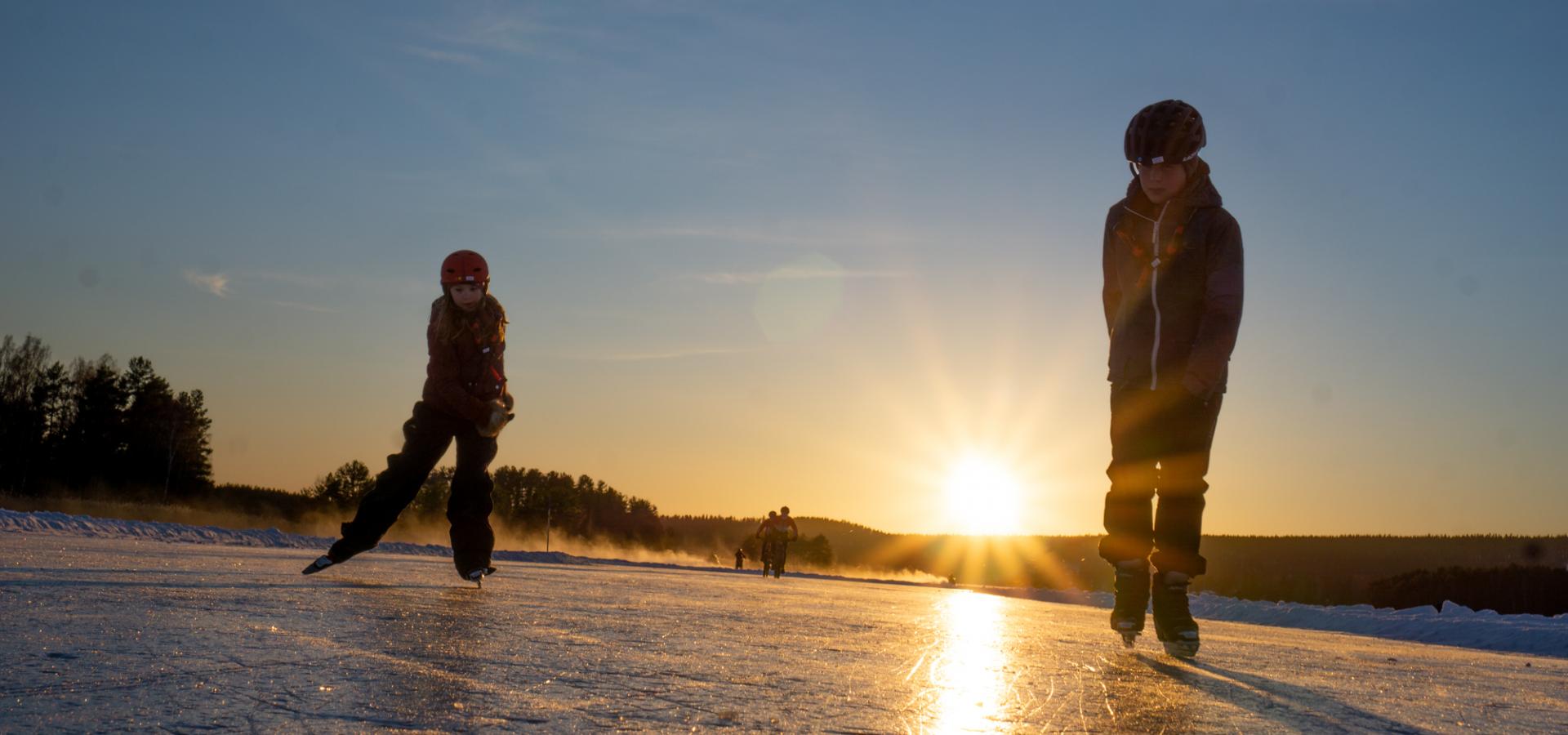 Två barn åker skridskor i solnedgången.