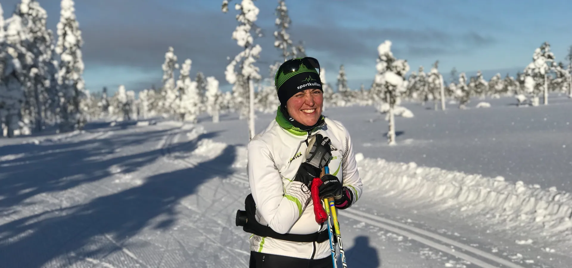 Marie Mörk i skidkläder lutar sig mot stavarna i soligt skidspår och ler.