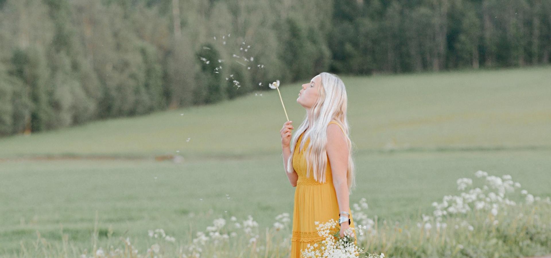Stina Jansson på en blomsteräng, blåser på en utblommad maskros.