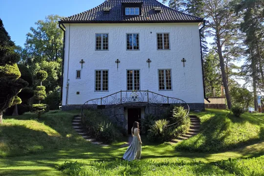 En kvinna går genöm en grönskande trädgård framför ett vitt hus sommartid.
