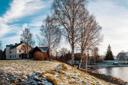Panoramavy över ett vitt hus och en vit kyrka, gräs, träd och vatten i förgrunden.