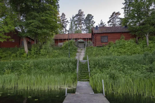 Faluröda äldre hus intill en sjö omgivna av grönska.