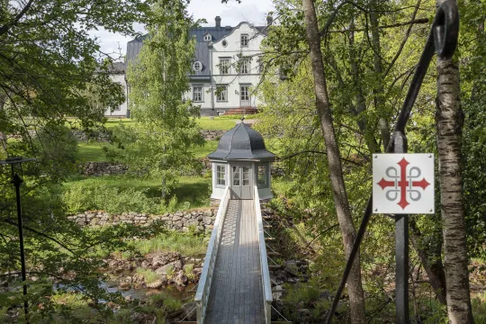 En träbro omgiven av grönska leder mot ett vitt hus.