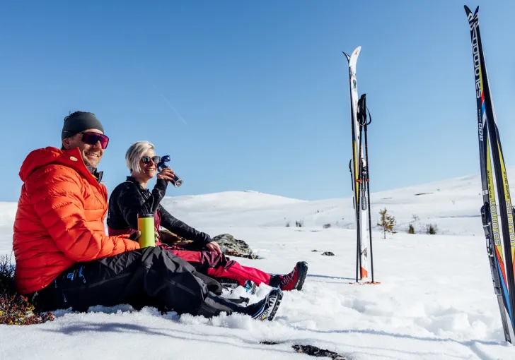 Ett par sitter och fikar i solen på fjället med skidorna nedstuckna i den djupa snön.