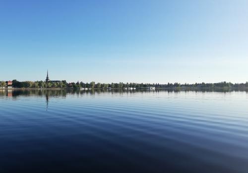 Blå himmel och blått vatten, träd och ett kyrktorn speglas i sjön.