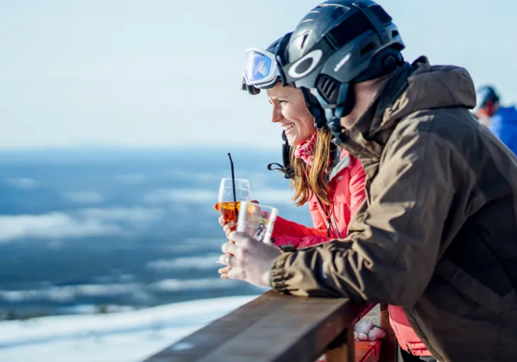 Ett par i slalomhjälmar håller i glas med dryck och lutar sig mot ett staket.