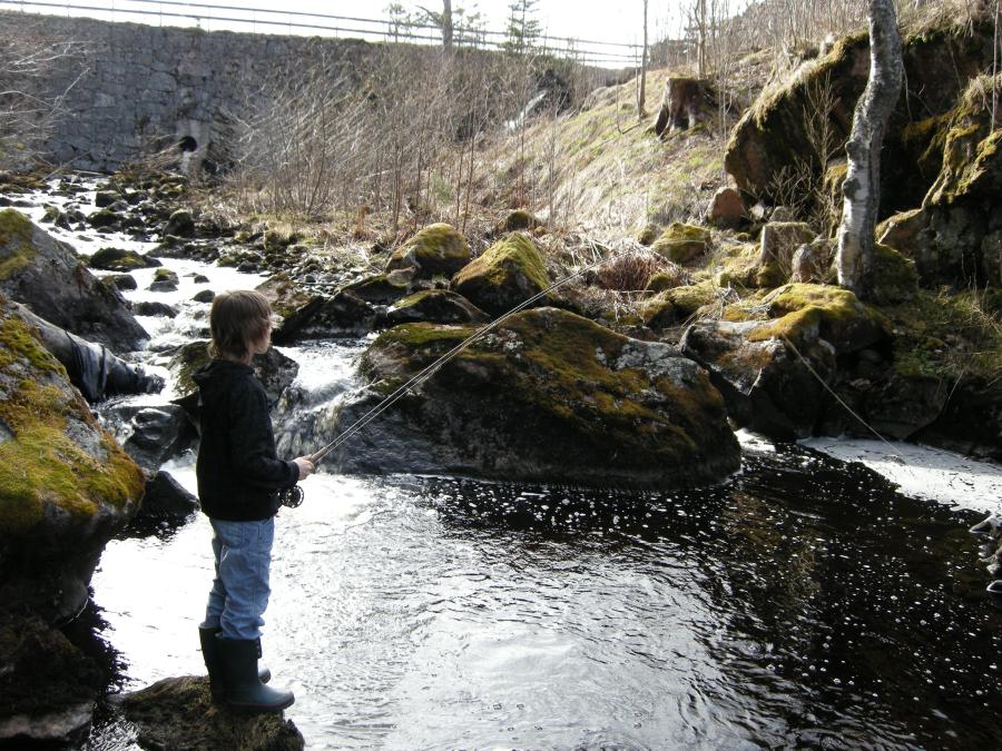 Pojke står och fiskar i bäck med med stora stenblock vid sidorna.