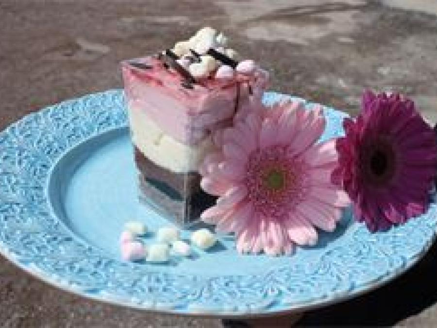 Ett bakverk i tre lager i brunt, vitt och rosa, visa minimashmallows och choklad på toppen, bakverket står på ett turkost keramikfat med spetskant, en rosa och en mörklila blomma ligger på fatet.