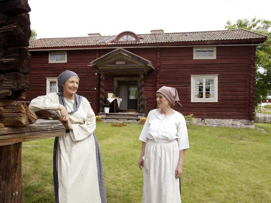 Två kvinnor i gammaldags bondeklädsel, en av dem lutar sig mot en husknut i timmer, en röd äldre timmerbyggnad i två våningar i bakgrunden.