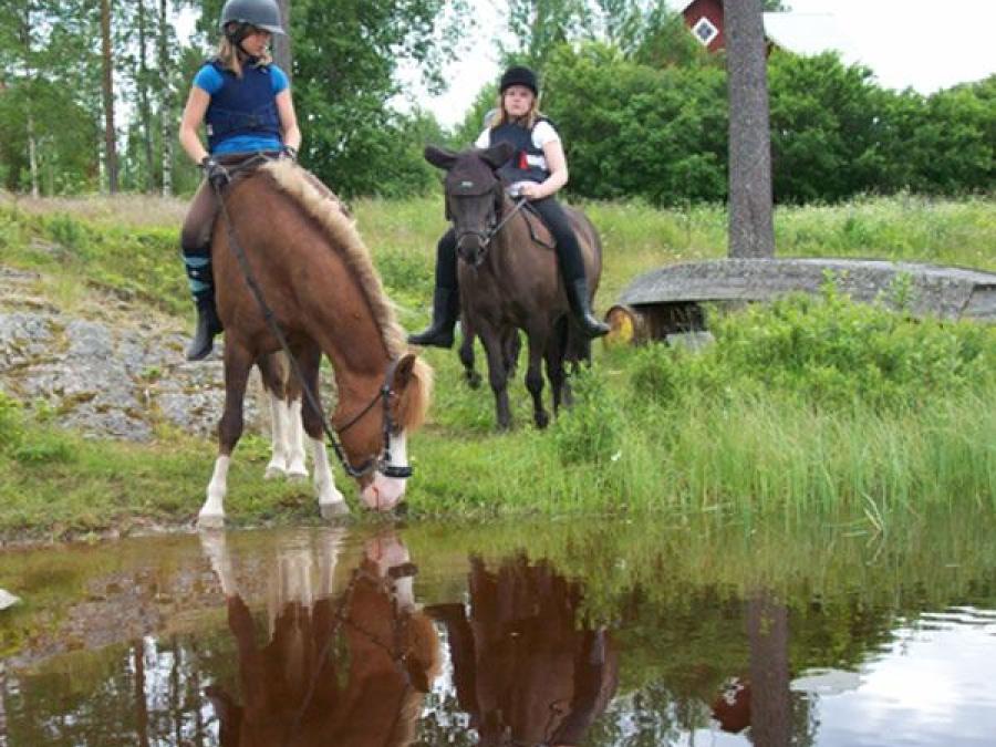 Två flickor på varsin häst, en av hästarna dricker vatten.