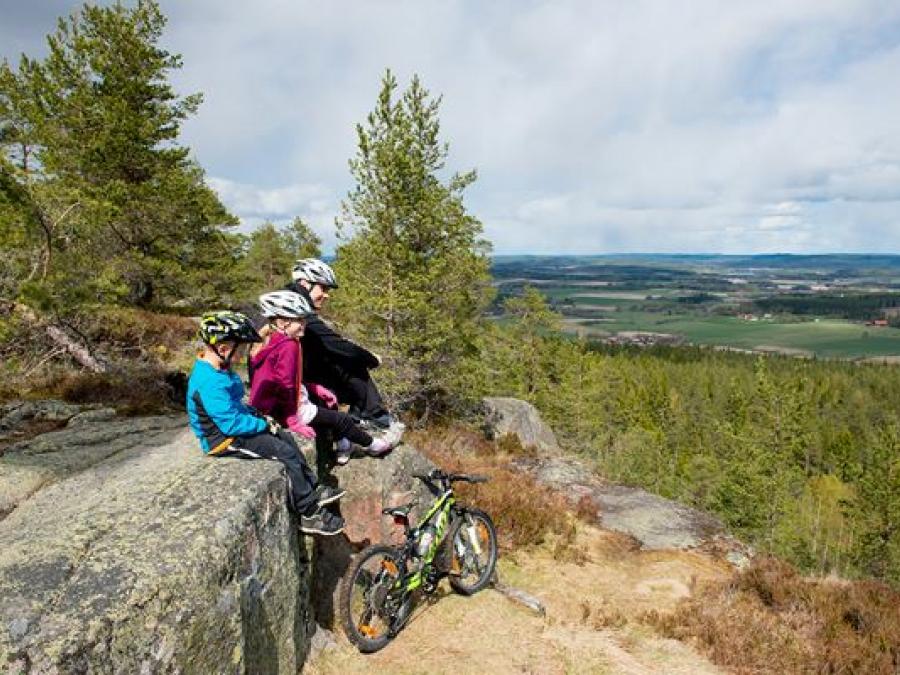En vuxen och två barn sitter på en liten klippa och tittar på utsikten, alla tre har cykelhjälm, nedanför klippan står en cykel.