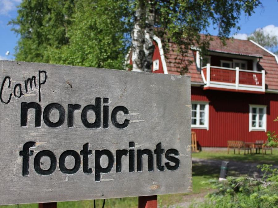 Skylt med camp nordic footprints med ett rödmålat hus i bakgrunden.