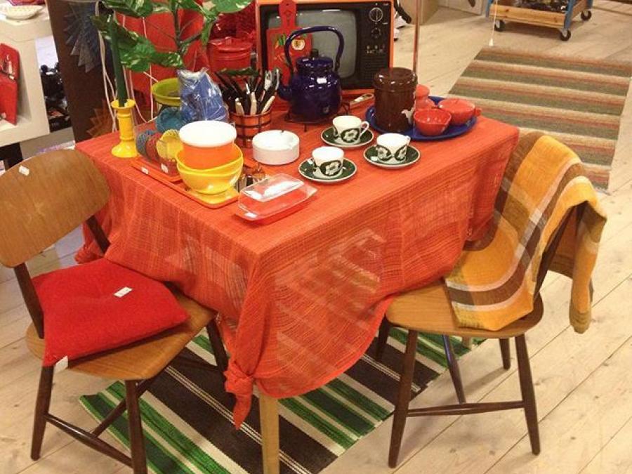 Interior image from the shop, a table with orange cloth, cups and other goods on the table, a striped rag rug on the floor.