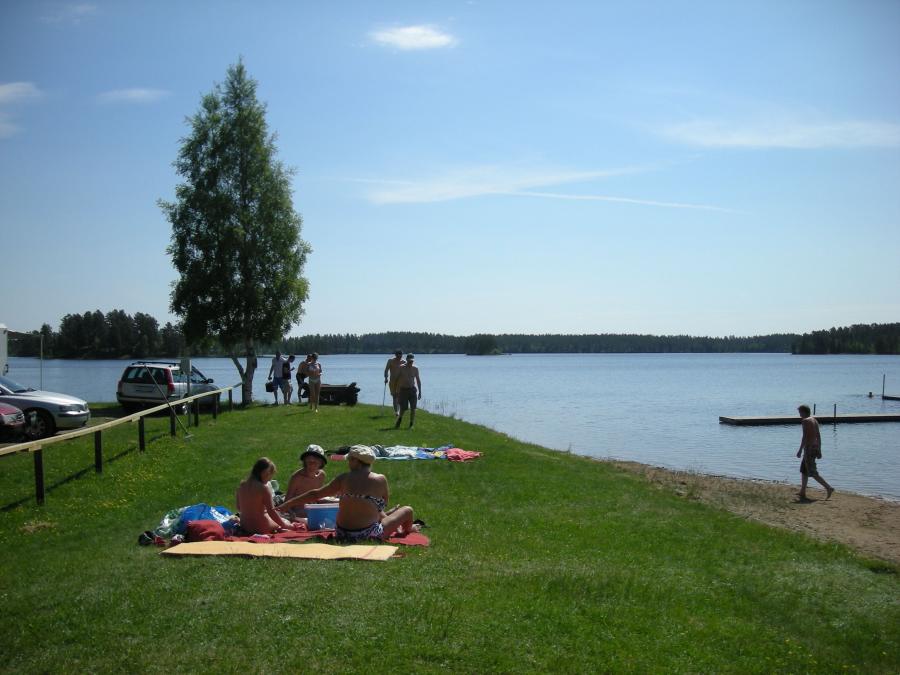 People sit on blanket by swimming lake with grass and sandy beach.