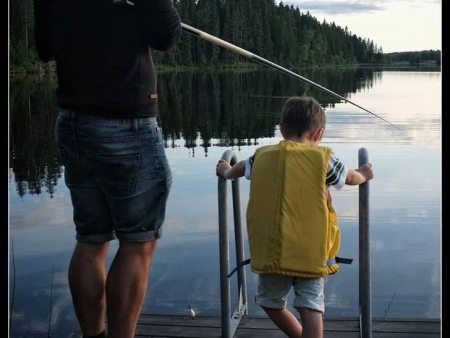 Pappa med fiskespö med son med flytväst på brygga vid sjö.