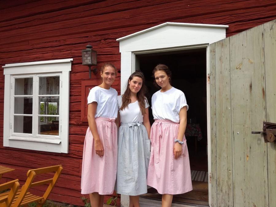 Tre tjejer med förkläden som står vid dörröppningen till bagarstugans cafe. 