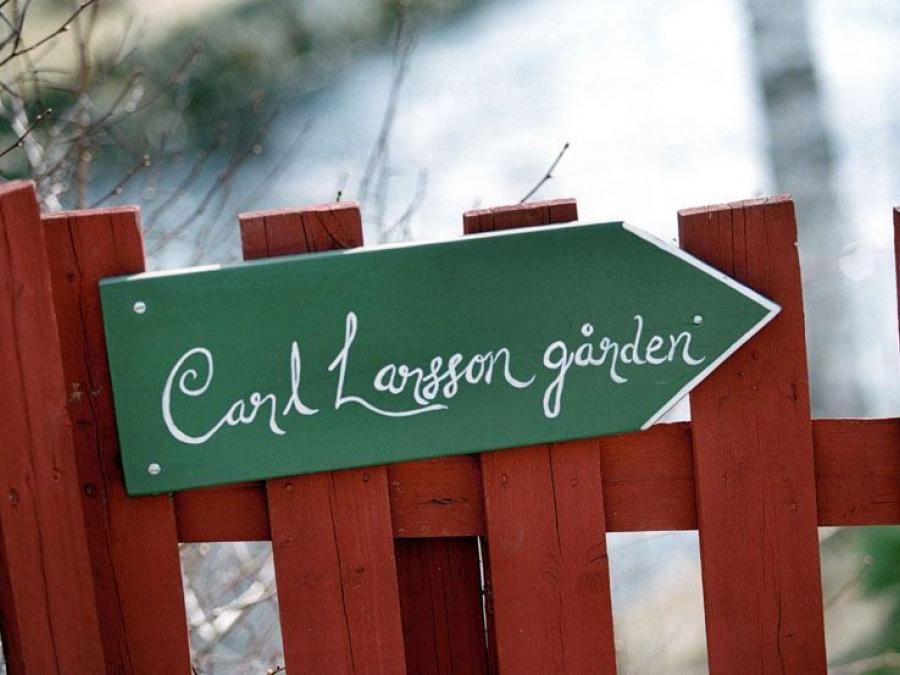 A green wooden sign with white text Carl Larsson gården, the sign is on a red wooden fence.