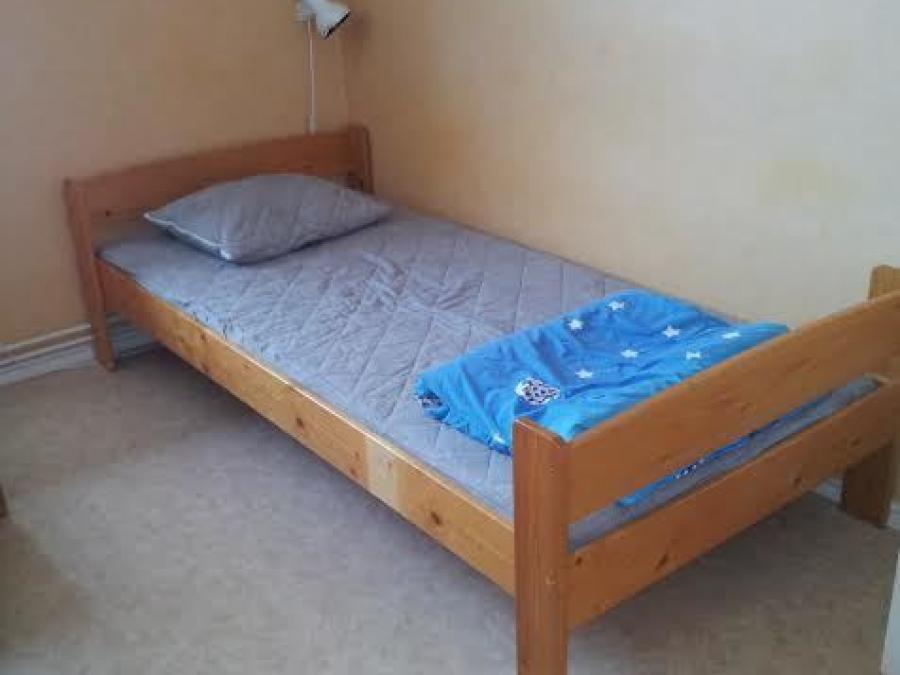 Enkelsäng med sängram i trä med ljusa väggar och ljusgrå plastmatta på golvet. 