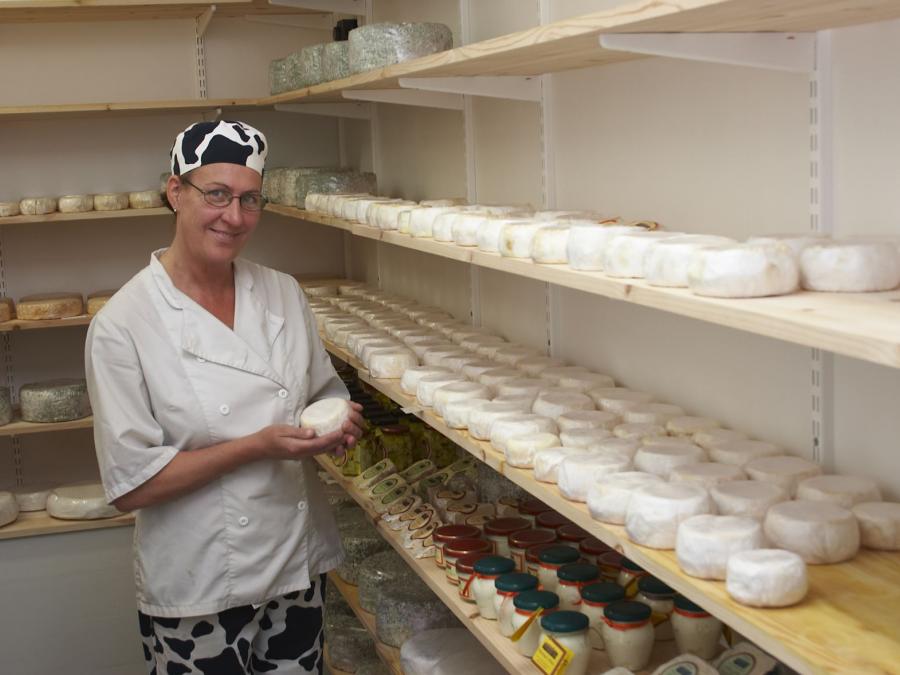 A woman in a white coat holding a cheese, cheeses lying on shelves around the room.