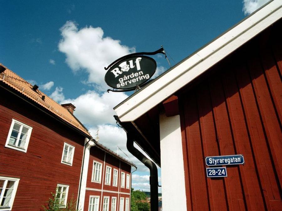 En röd husknut, på väggen en blå skylt Styraregatan, på taket en svart skylt Rolfgården servering, röda trähus på andre sidan gatan.