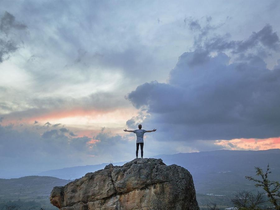 En person står högst upp på en stor sten med armarna utsträckta och tittar på utsikten.