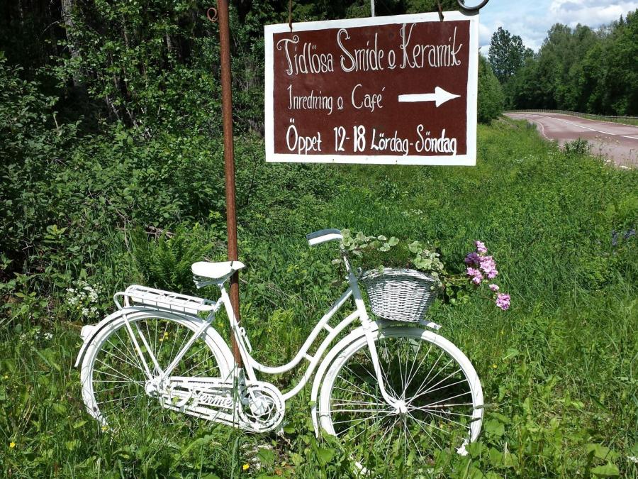 A white women's bicycle standing in the grass, above the bicycle hangs a sign with the text Tidlösa smide & keramik opening hours.
