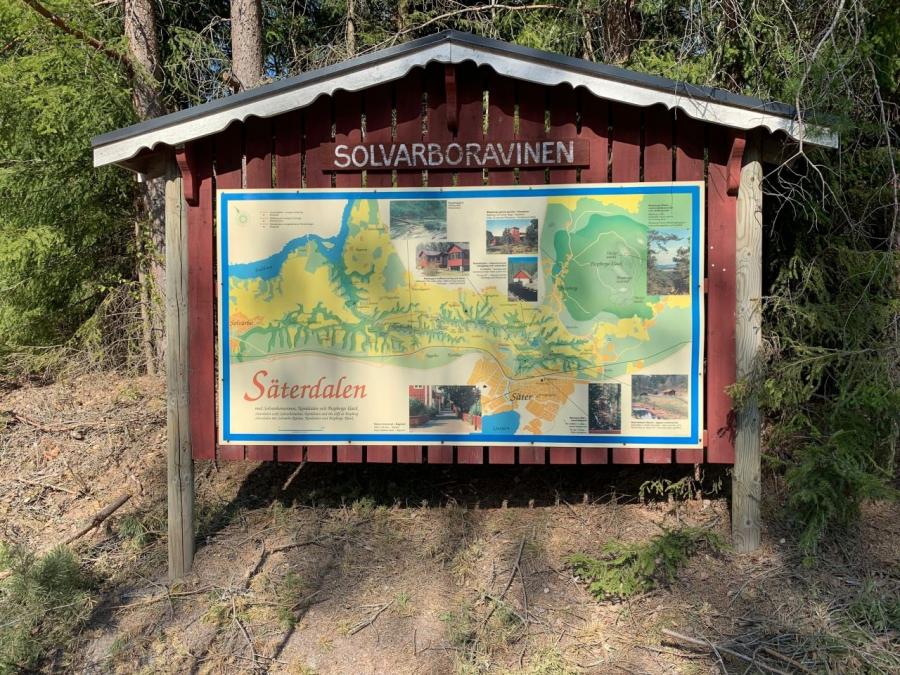 Informationstavla med en karta som visar Solvarboravinen och beskrivande text, tavlan är i rött trä, träd i bakgrunden.